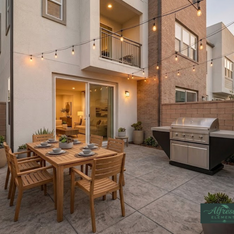 Outdoor patio with wooden dining table and chairs, grill, and string lights.