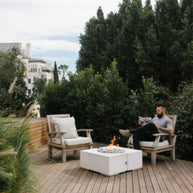 Person sitting on a wooden deck with chairs and a fire pit, surrounded by greenery.