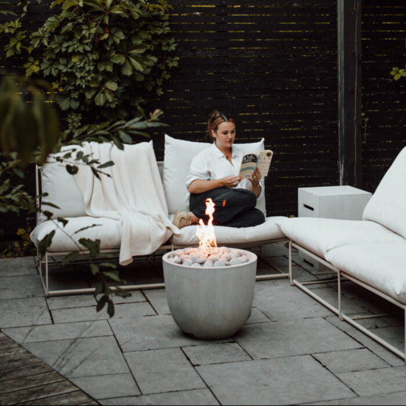 Woman reading a book on a patio with a fire pit and white outdoor furniture.