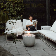Woman reading a book on a patio with a fire pit and white outdoor furniture.