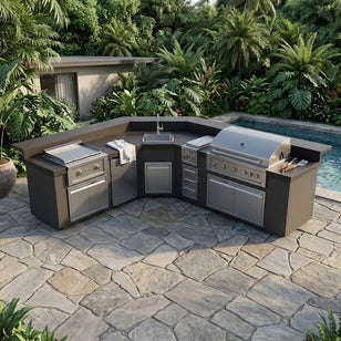 Outdoor kitchen with grill and sink area near a pool, surrounded by lush greenery.
