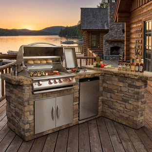 Outdoor kitchen with grill and cabinets on a wooden deck by a lake at sunset.